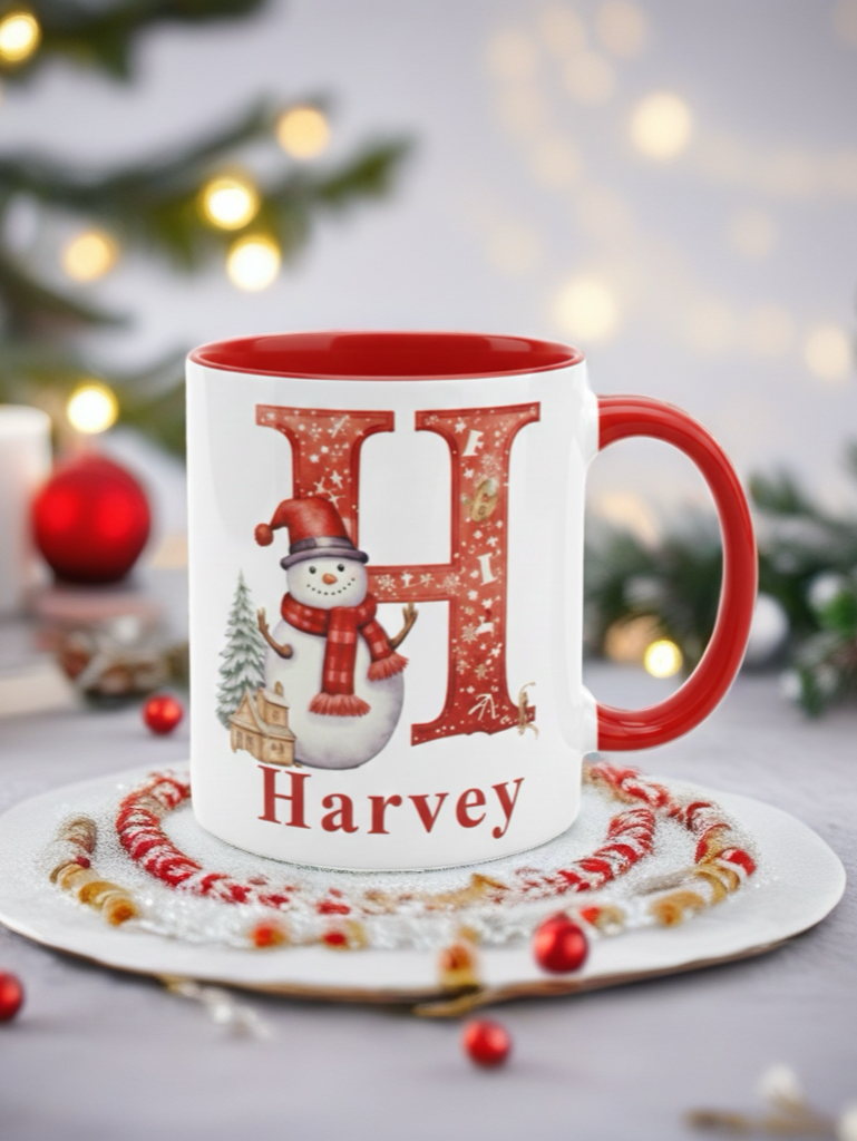 A white ceramic mug with a red interior and handle, decorated with a large glittery red letter H, a snowman, a small tree, and the name Harvey. The mug sits on a white plate with red and gold beaded trim, with a softly blurred holiday background featuring lights and greenery.